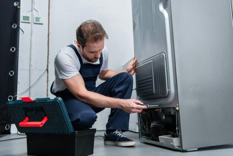 adult bearded repairman in working overall fixing refrigerator by screwdriver in kitchen